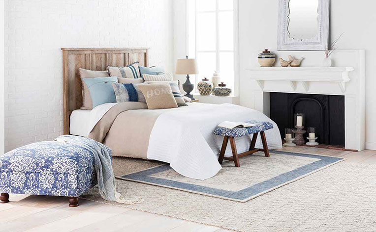 Bedroom with a bed draped in tan and white bedspread covered in square throw pillows and surrounded by round vases.